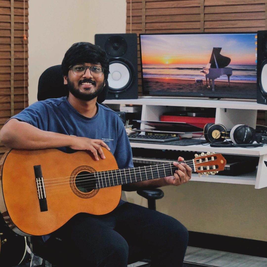 Man playing guitar in a home studio with music equipment and a screen displaying a piano on a beach.